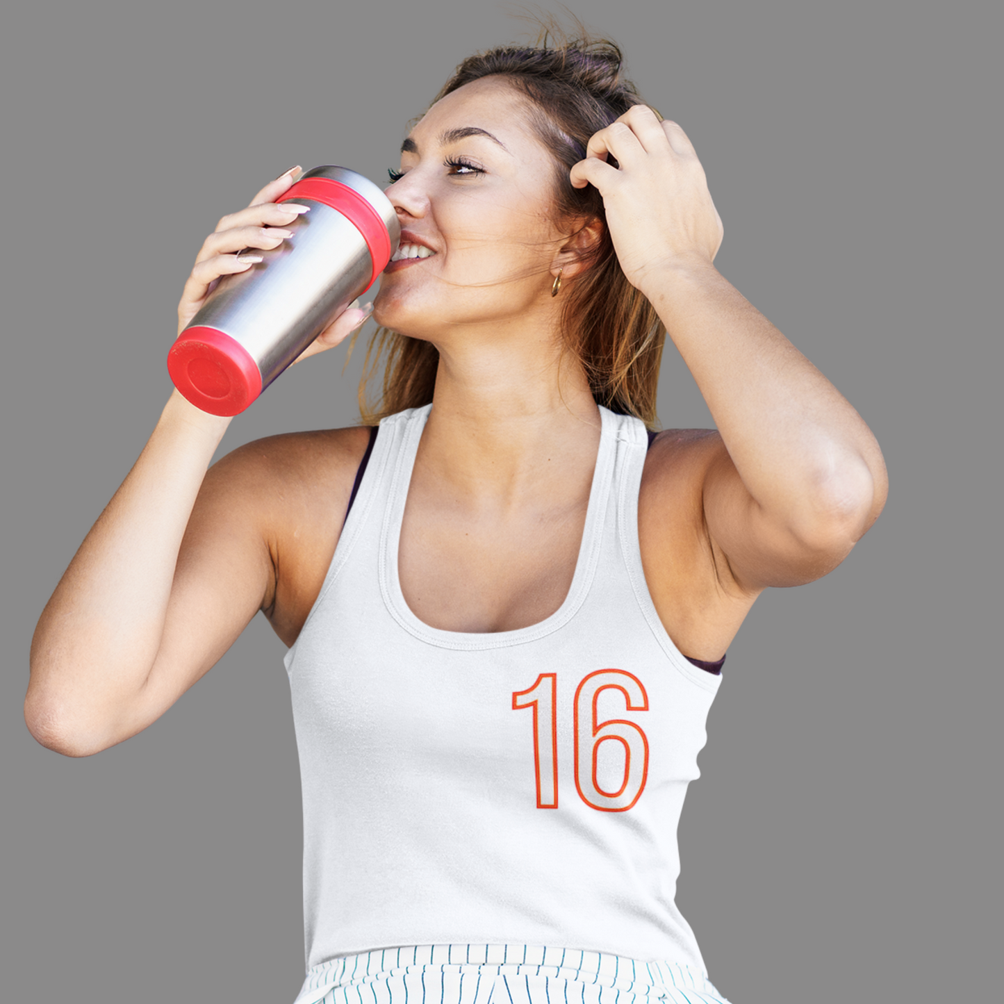 Woman in a white tank top with the number 16 drinking from a silver and red water bottle on a gray background