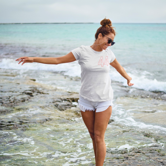 Urban Rush Originals Women's 44 Quali Day Relaxed Soft Tee in White, worn by a woman with sunglasses and white shorts balancing on rocks at the beach, showcasing a comfortable and relaxed fit.