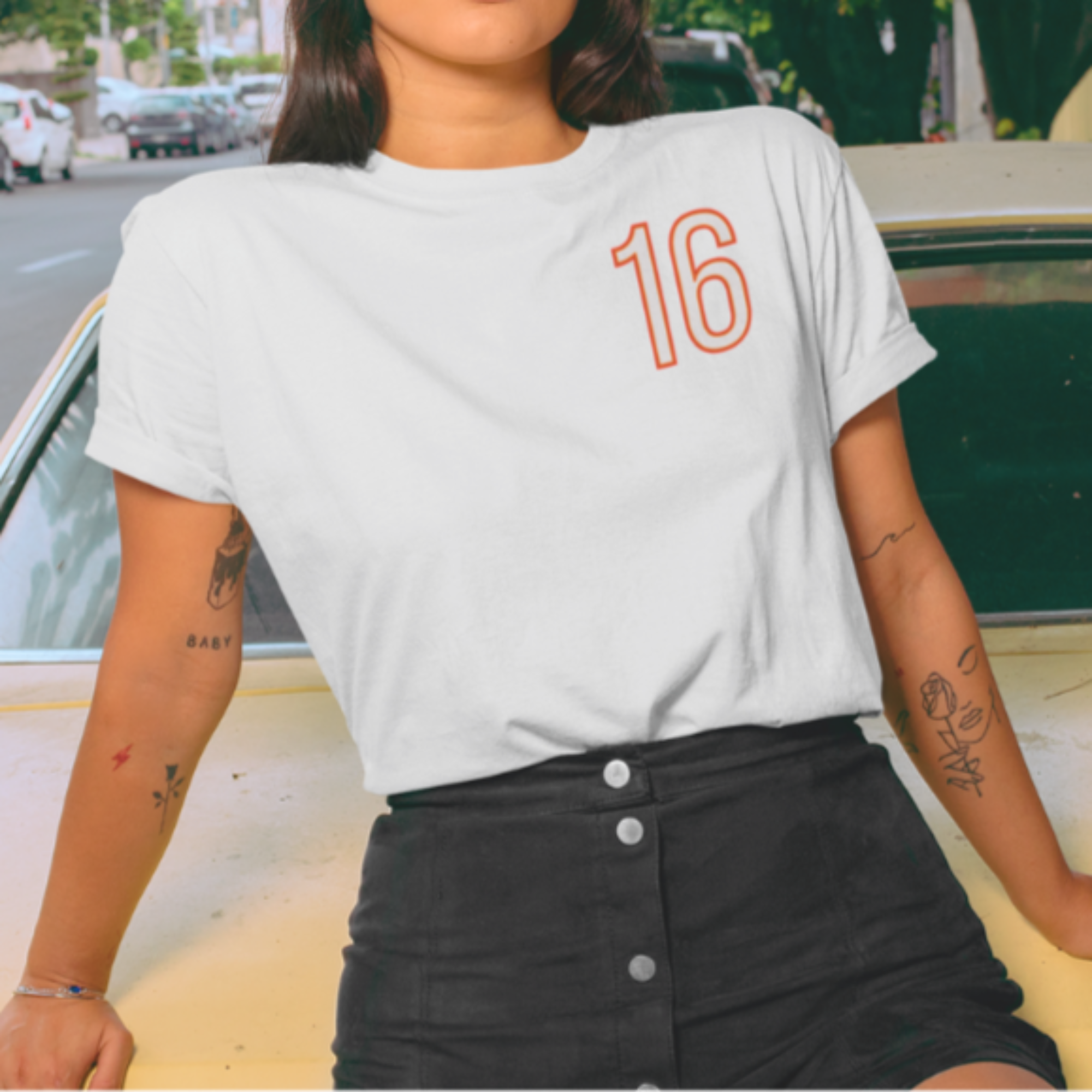 Person wearing a white t-shirt with the number 16, standing next to a vintage car on a street.