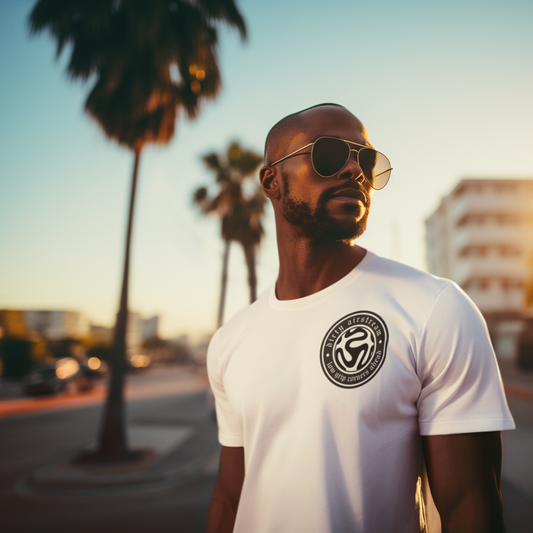 Man wearing a white t-shirt with a logo, sunglasses, and a beard, standing on a street with palm trees and buildings in the background.
