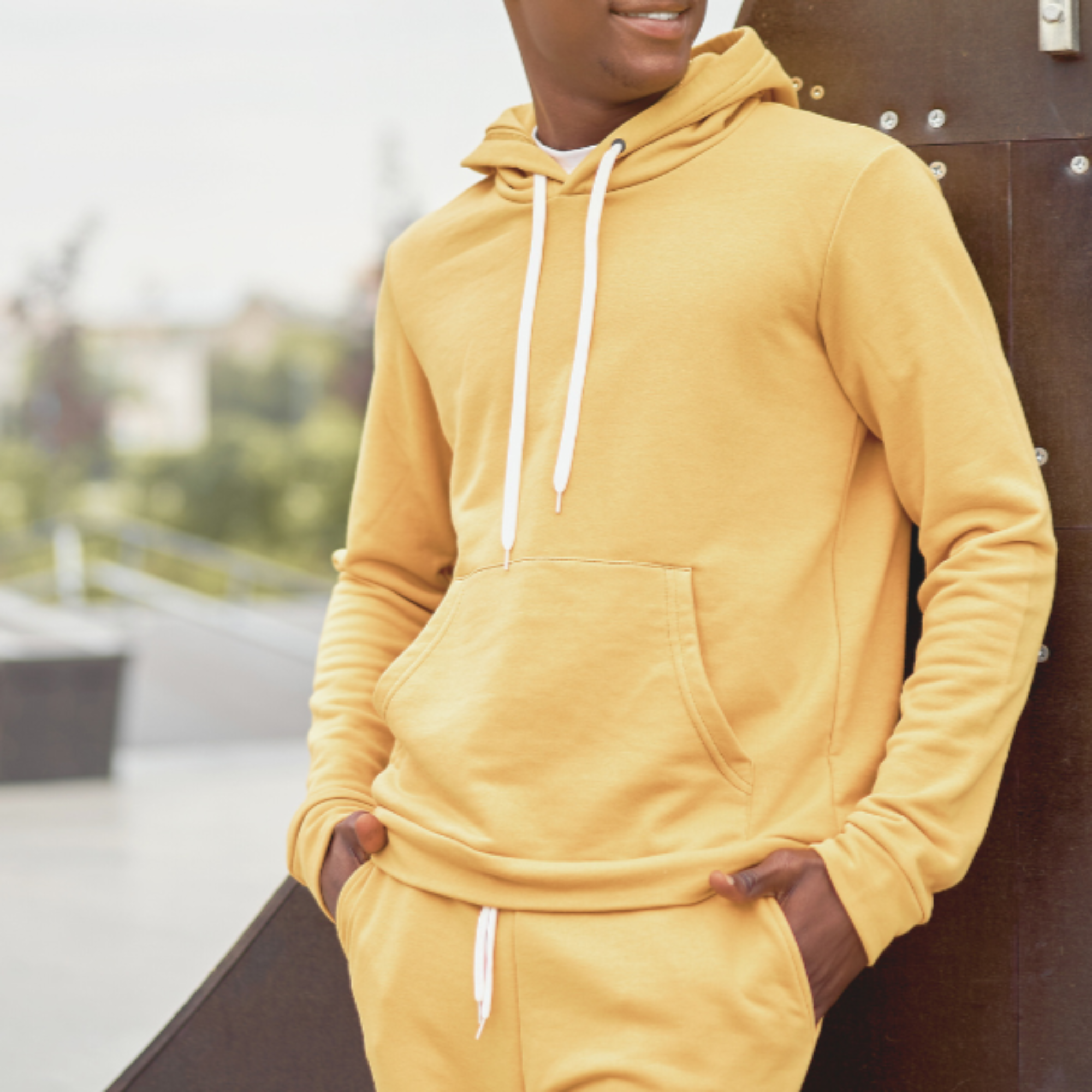 Person wearing a mustard yellow hoodie and pants leaning against a wall with a blurred outdoor background.